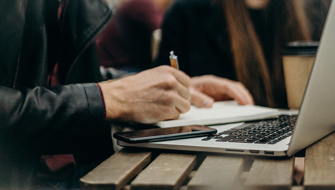 Imagem destacada do artigo "Como ser perito judicial em 2023 - paga bem?" Blog Perícia Expert, do professor André Bittencourt. Na imagem temos a mão de um homem escrevendo em uma agenda por cima de um laptop, com um celular do lado