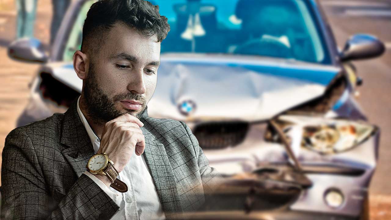 A imagem contém no primeiro plano um homem adulto, usando roupa social, olhando para baixo, com a mão direita no queixo e semblante pensativo. Ao fundo da imagem a frente de um carro batido desfocado.