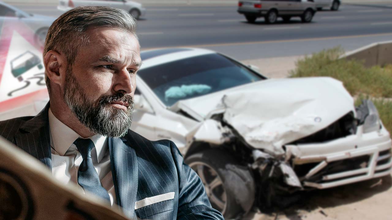 A imagem contém no primeiro plano a parte superior de um homem, com barba, vestindo terno, olhando para o lado. Ao fundo, desfocado, um carro branco acidentado na estrada, e uma placa de transito desfocada.